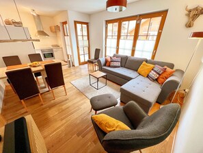 Ein modernes Wohnzimmer mit grauem Sofa, einem Esstisch mit Stühlen, einer Küchenzeile und großen Fenstern mit Jalousien.