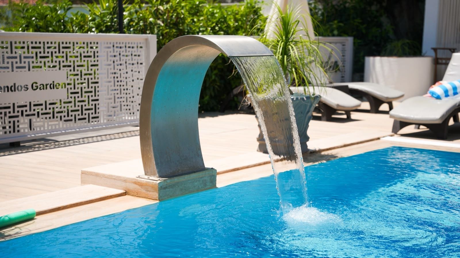 A swimming pool with a modern water feature, surrounded by lounge chairs and a patterned fence.