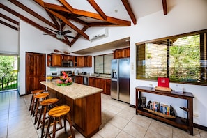 A kitchen with wooden cabinets, a central island with stools, a refrigerator, and a window with blinds.