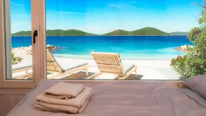 Ein Zimmer mit großem Fenster und Blick auf einen Strand, zwei Liegestühle und ein ordentlich bezogenes Bett.