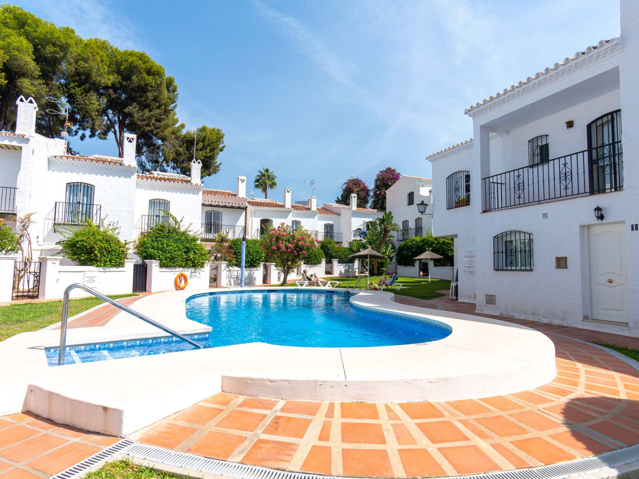 Piscina rodeada por edifícios brancos com varandas, céu claro e um jardim bem cuidado.