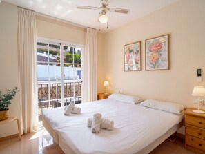 Une chambre d’hôtel avec un grand lit, une table de chevet, une commode, une lampe, un ventilateur de plafond et deux tableaux floraux encadrés.