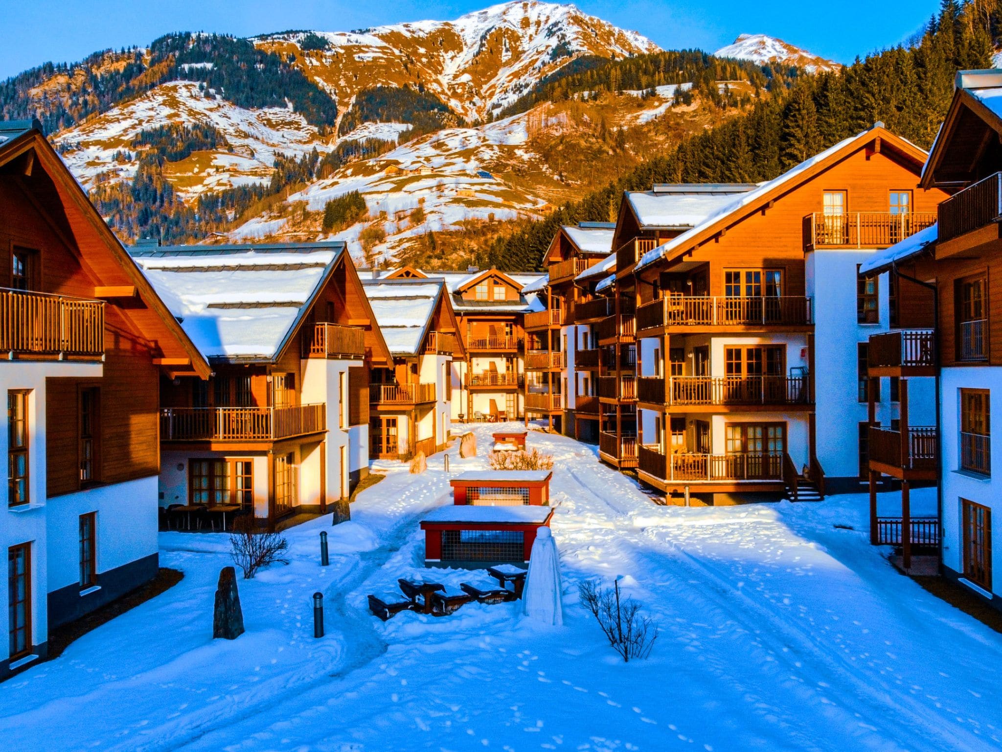 A snow-covered village with wooden buildings, a central bench, and mountains in the background.