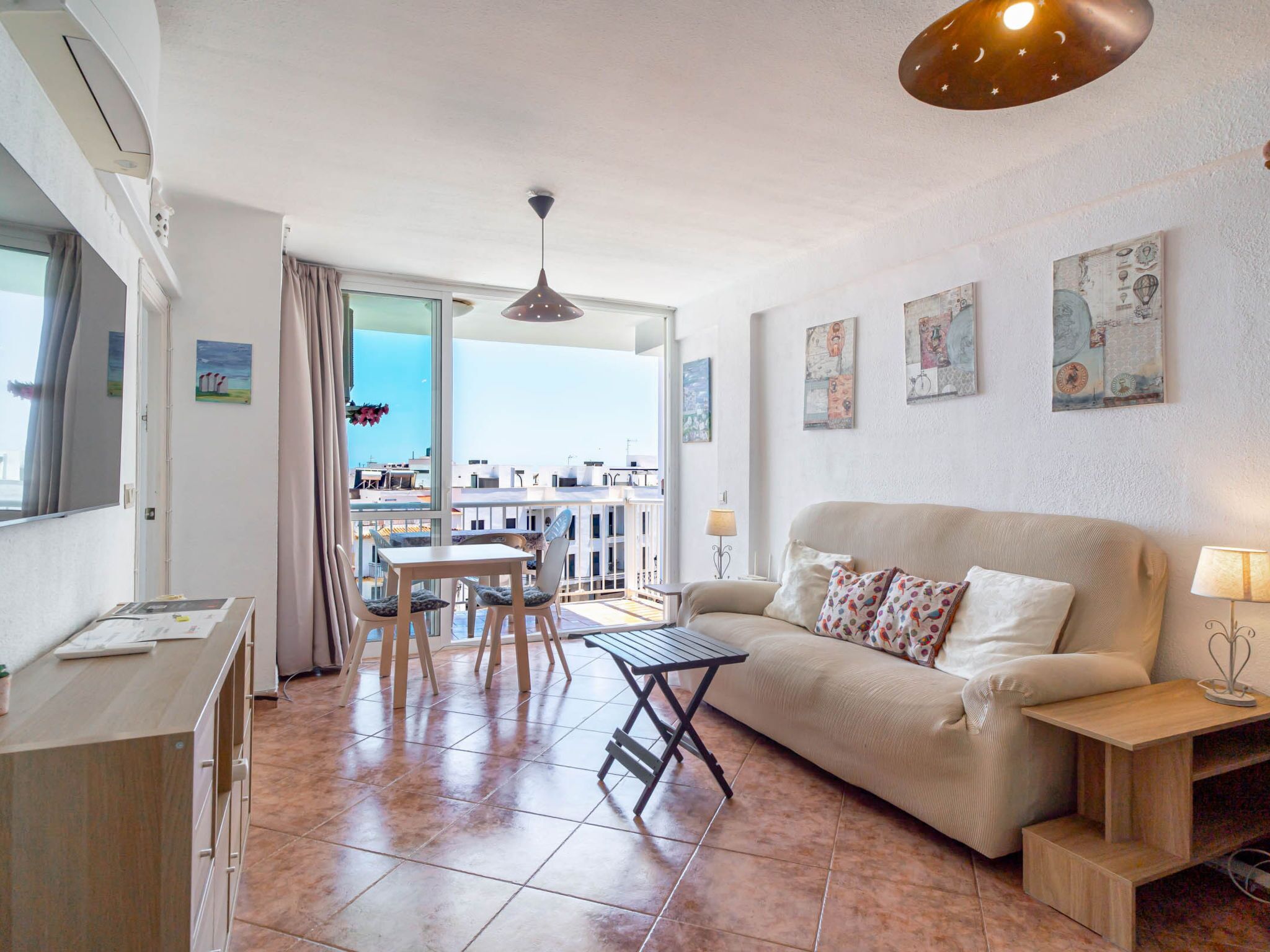 A modern living room with a sofa, a dining table, and chairs. There is a balcony with a view of buildings.