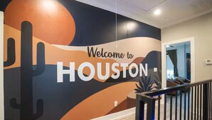 En væg med et 'Welcome to Houston'-kunstværk, en trappe og udsigt ind til et rum med en seng og et vindue.