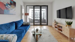 A modern living room with a blue sofa, a glass coffee table, a wooden TV stand, and a large TV. - Plac Unii by Grand Apartments (Gdynia)