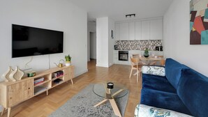A modern living room with a flat-screen TV, wooden console, blue sofa, and a kitchen in the background. - Plac Unii by Grand Apartments (Gdynia)
