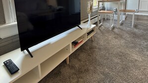 A modern living room with a large flat-screen TV on a white entertainment unit, a kitchen area with white cabinets, and a view of greenery outside. - Cozy Private 1BR in the Heart of 5 Points (Denver)