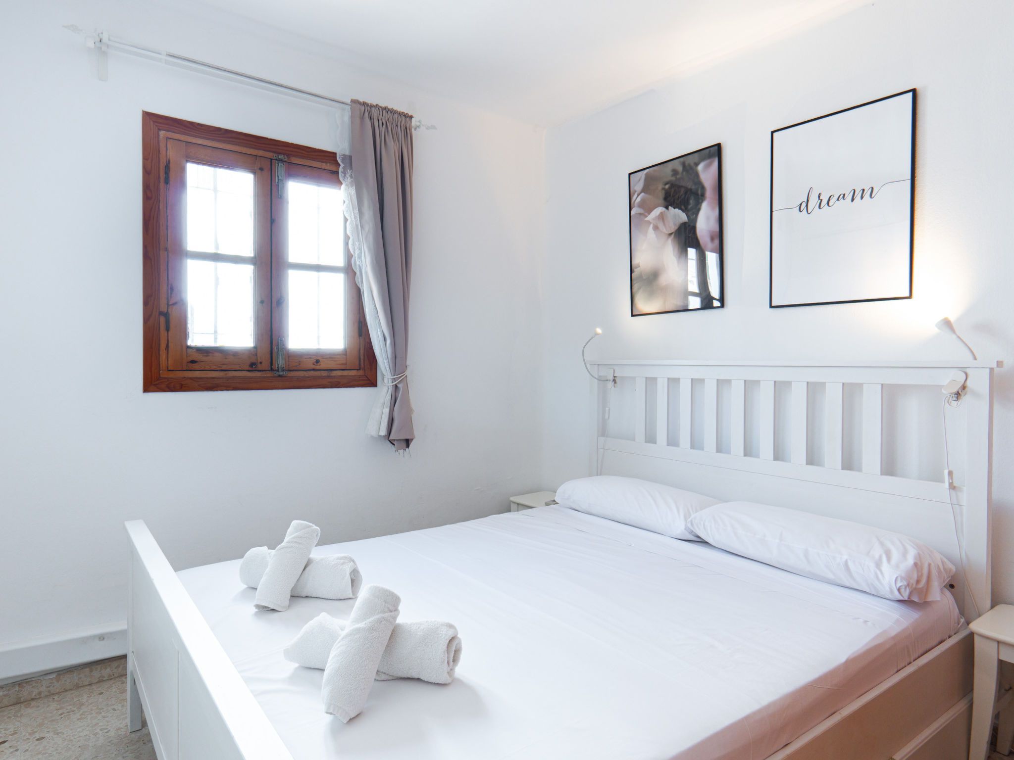 Un dormitorio con cama blanca, cabecero de madera, ventana con cortinas y dos cuadros en la pared.