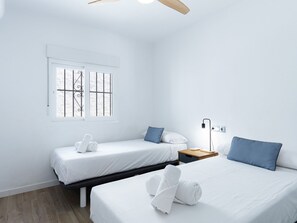 Ein Hotelzimmer mit zwei Betten, einem Fenster mit Gitterstäben, einem Nachttisch mit Lampe und einem Deckenventilator.