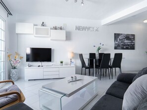 Ein modernes Wohnzimmer mit Flachbildfernseher, einem Glastisch, einem Essbereich mit weißem Tisch und schwarzen Stühlen sowie einer Wandregal mit Dekorationselementen.