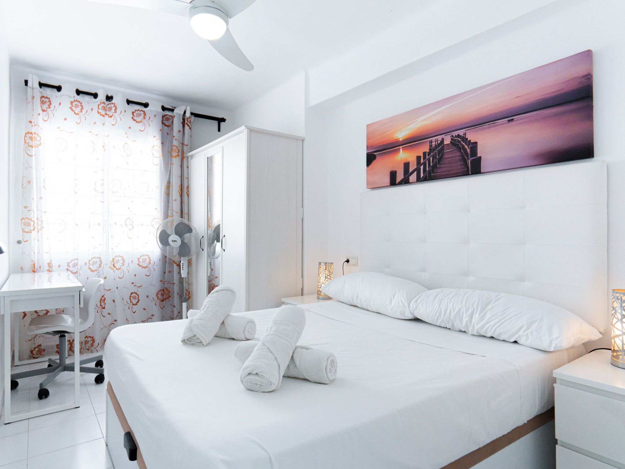 Une chambre d’hôtel avec un lit, un bureau, une chaise, un ventilateur sur pied et un tableau représentant un coucher de soleil.