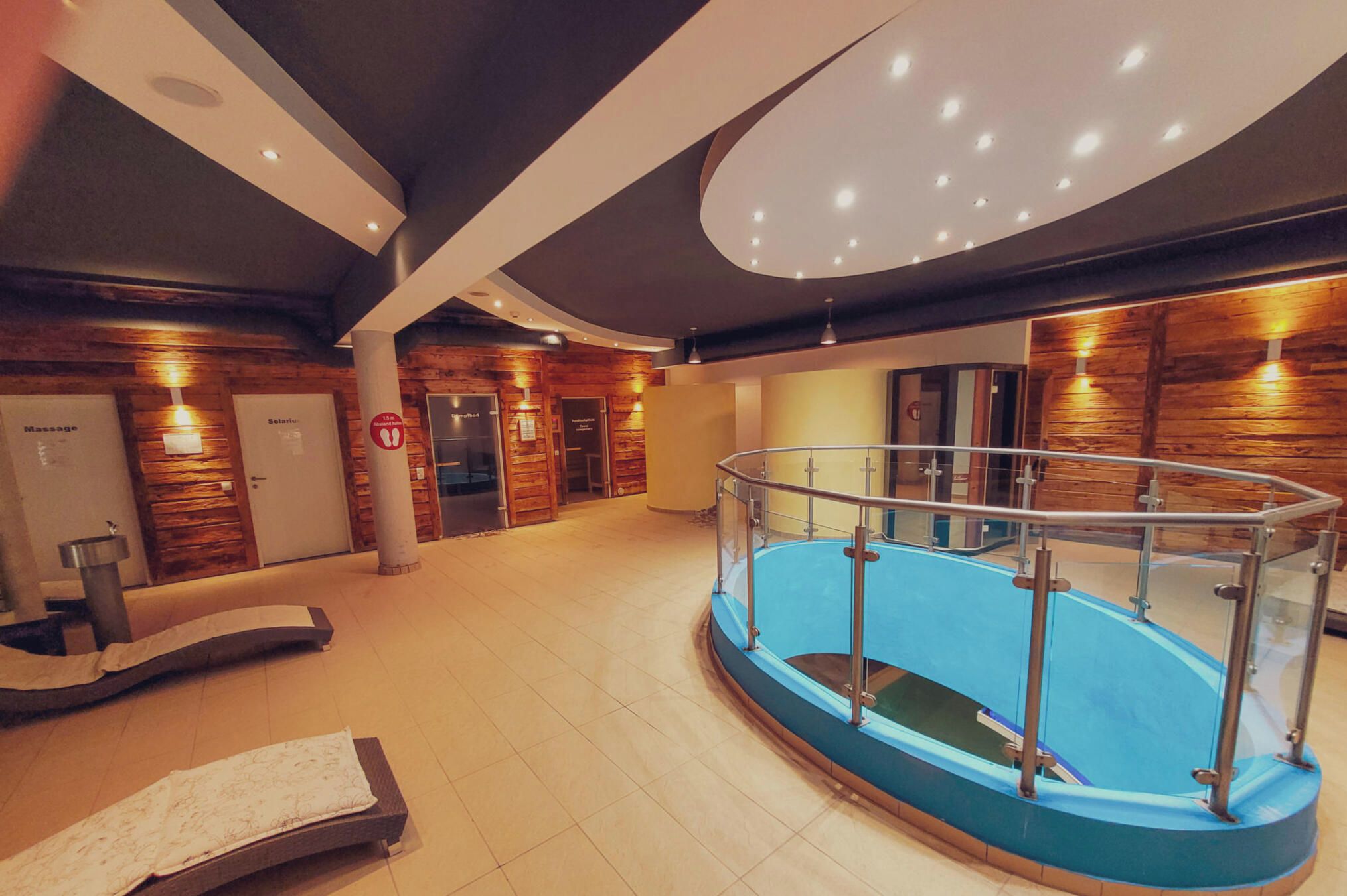Zona d'una piscina interior moderna amb barana de vidre, parets de fusta i una sala de massatges a l'esquerra.
