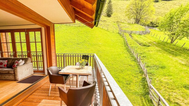 Un terrazzo in legno con un divano e sedie, un tavolo con un vaso di fiori, e vista su una valle verde.