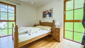 Una camera da letto con un letto in legno, due comodini, un tavolino e un quadro, con ampie finestre che offrono vista su un paesaggio verde.