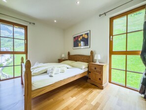 Ein Schlafzimmer mit einem hölzernen Bett, zwei Nachttischen, einem kleinen Tischchen, einem Gemälde und großen Fenstern mit Blick auf eine grüne Landschaft.