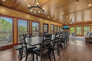 A spacious dining area with a long table and chairs, wooden ceiling, and large windows with blinds.