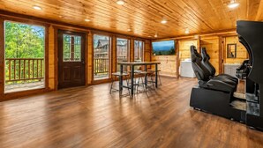 A spacious room with wooden floors, a dining area with a table and chairs, a large TV mounted on the wall, and a seating area with a sofa and a massage chair. - Sleeps 20 | Private Indoor Pool, Theater, Arcade (Sevierville)