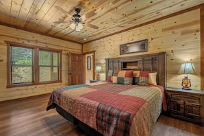 A log cabin bedroom with a bed, wooden walls, a ceiling fan, and a window with blinds.