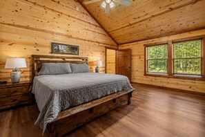 Ein Schlafzimmer einer Holzhütte mit einem Bett, Nachttischen, einem Deckenventilator und einem Fenster mit Blick auf Grünflächen.