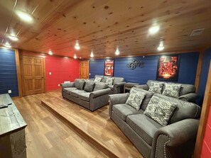Quarto com piso de madeira, parede vermelha e azul, e teto com iluminação embutida. Há dois sofás cinzas com almofadas estampadas.
