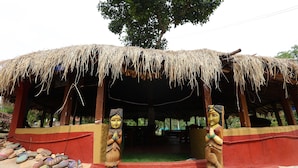 A thatched-roof structure with two wooden statues, a red and yellow painted wall, and a green carpeted area. - Sakleshpur Calm Stay (Alur)