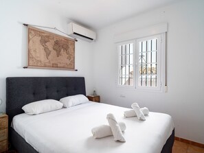 Une chambre à coucher avec un grand lit, du linge de lit blanc, une carte du monde accrochée au mur, un climatiseur, une fenêtre avec des barreaux et une table de chevet avec une lampe.
