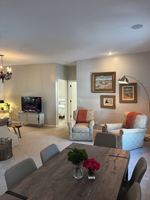 A modern living room with a dining table, two patterned armchairs, a television, and framed artwork on the wall.