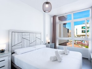 A hotel room with a bed, a nightstand, a lamp, a window with a view of buildings, and a hanging light fixture. - Apartment  by Interhome (Nerja)