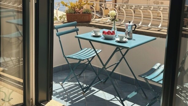 A balcony with a table set for two, a potted plant, and a view of the sea and a building with a dome. - De Vivo Realty - The Colombo Positano (Positano)