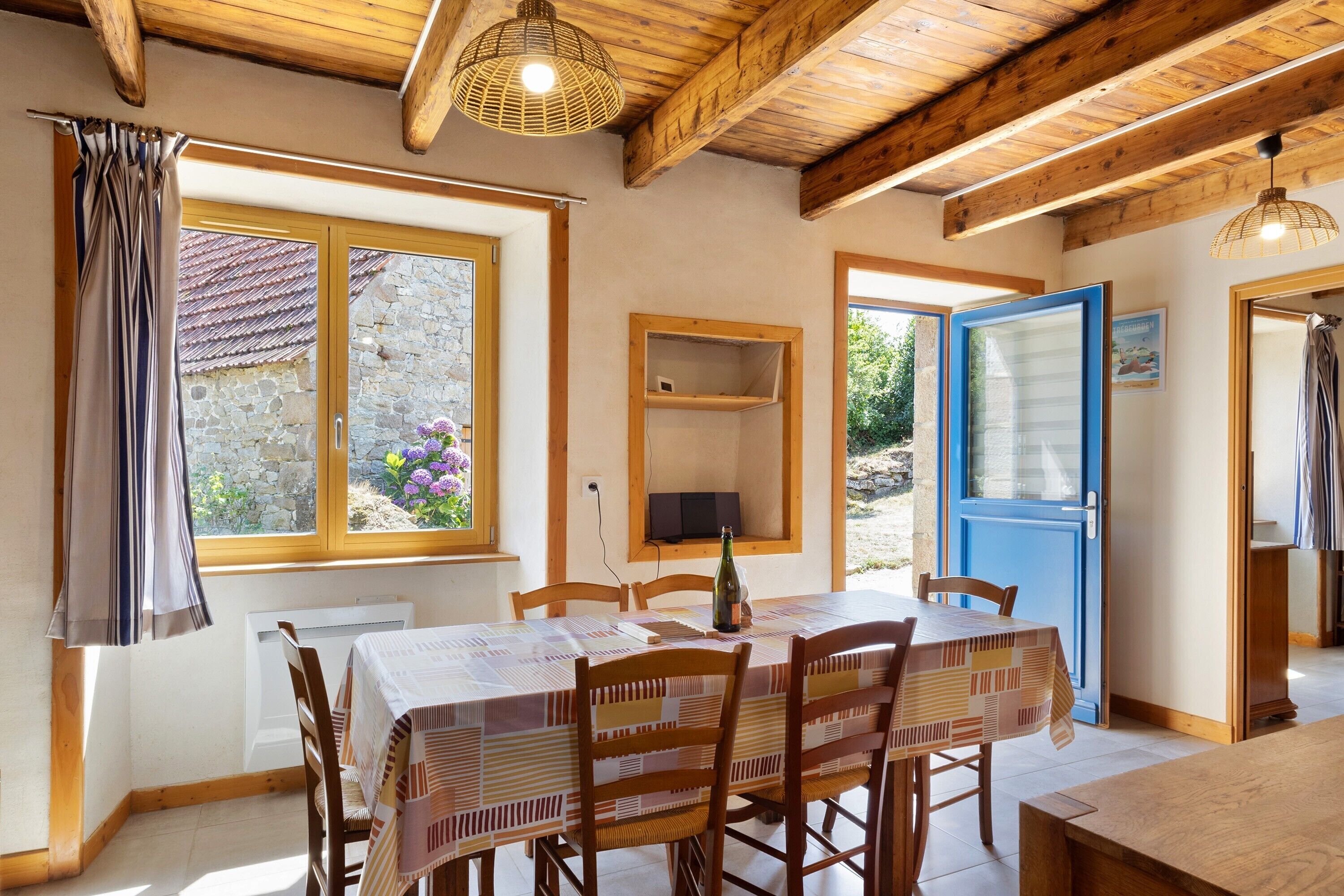A dining area with a table set for four, wooden chairs, a window with curtains, and a view of a stone wall and greenery outside.