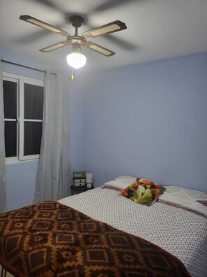 Une chambre à coucher avec un lit, un ventilateur de plafond, une fenêtre avec des rideaux et une table de chevet avec une lampe.