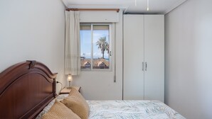 Une chambre à coucher avec une tête de lit en bois, un lit recouvert d’une literie à motifs, une table de chevet avec une lampe, une fenêtre donnant sur des immeubles et des palmiers, et une armoire.