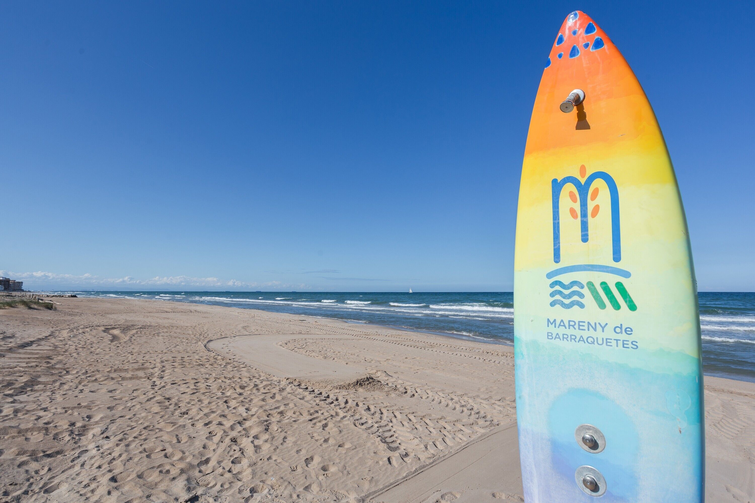 Una tabla de surf colorida con el texto 'Mareny de Barraquetes' en una playa, con huellas que conducen al agua.