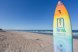 Une planche de surf colorée, portant l’inscription « Mareny de Barraquetes », sur une plage où l’on peut voir des empreintes de pas menant à l’eau.
