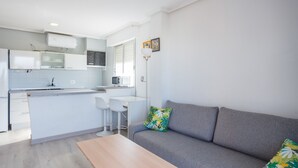 Une cuisine moderne avec des meubles blancs, un coin repas doté d’une table blanche et un canapé gris agrémenté de coussins aux motifs tropicaux.