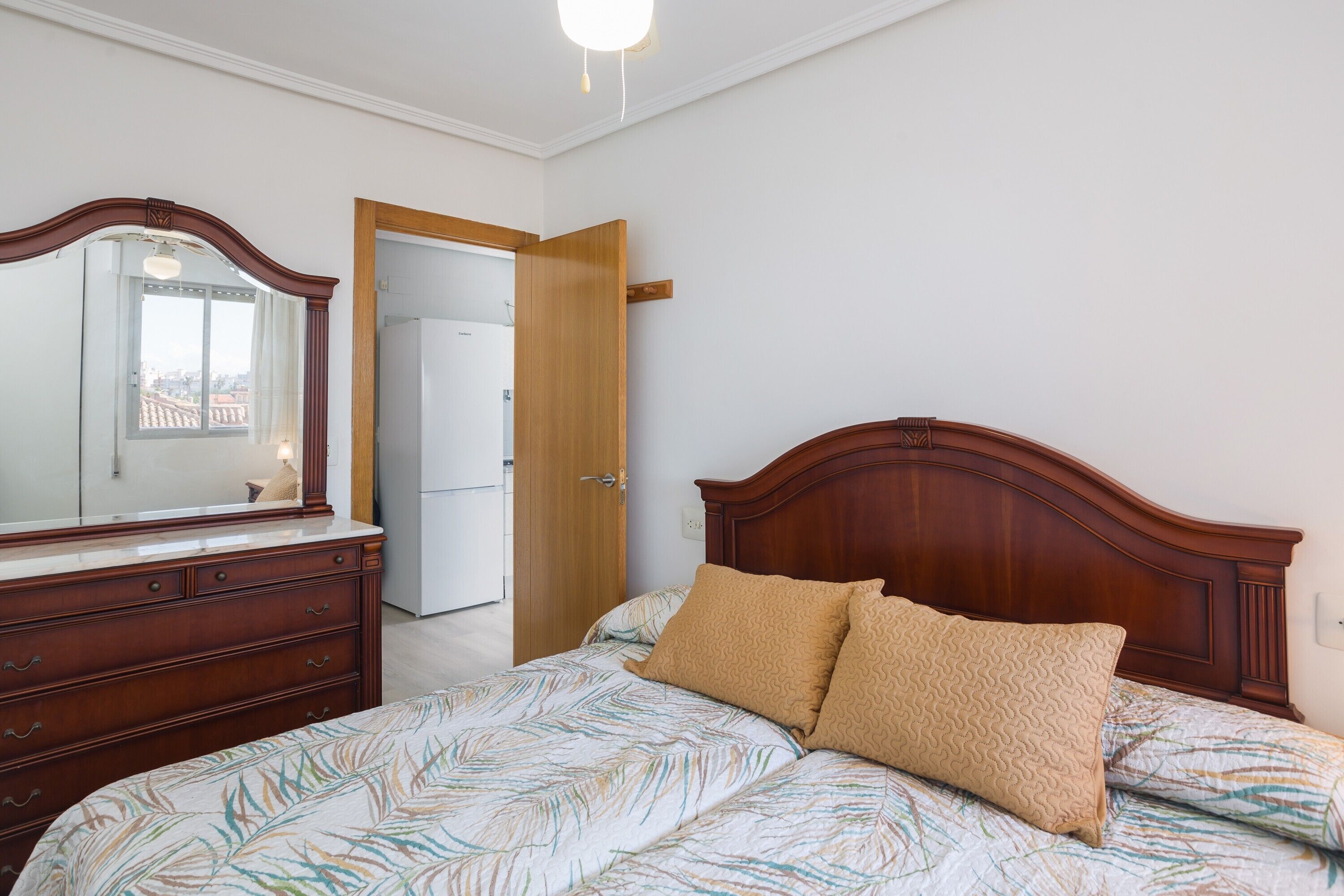 Een slaapkamer met een houten bed, een kapstok, een spiegel en een deur die naar een andere kamer leidt.
