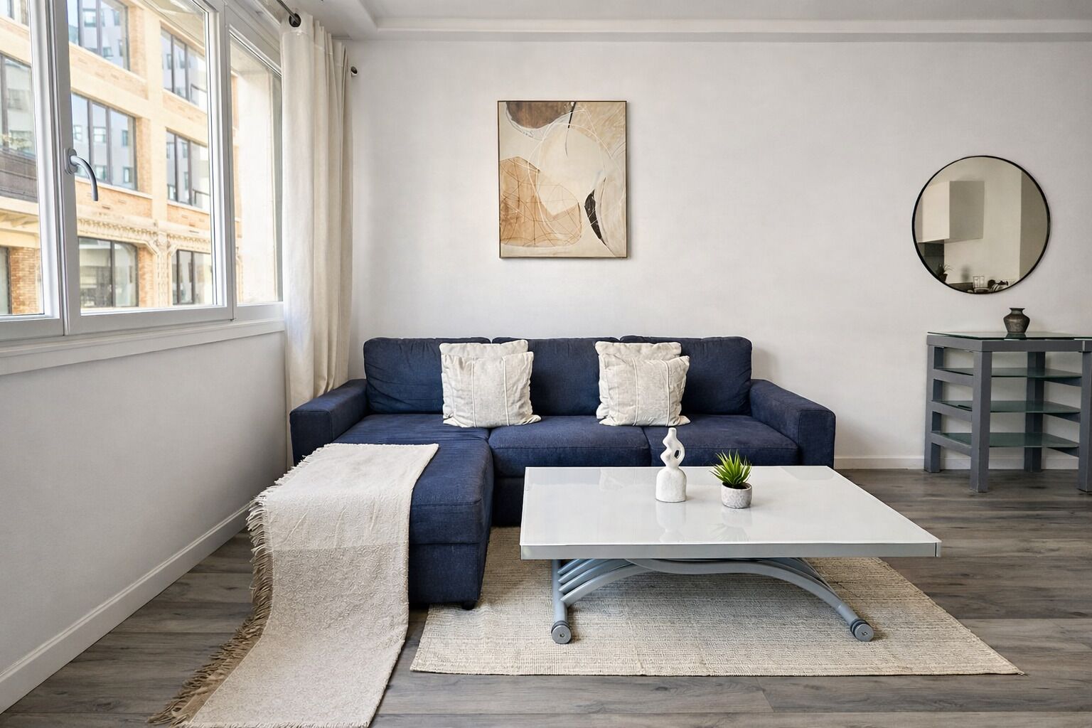 Un salon moderne avec un canapé d’angle, une table basse blanche, une petite plante, un tapis et un tableau accroché au mur.