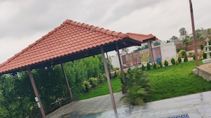 A swimming pool with a tiled edge, a covered area, and a grassy area with a fence and trees in the background.