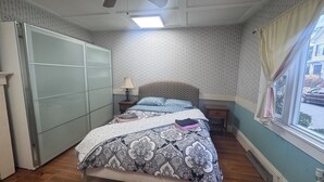 A bedroom with a bed, a nightstand, a dresser, a window with curtains, and a ceiling fan. - Inviting 1BR Near Boston -- Easy Transit -- Parking Included (Malden)