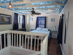 Ein Schlafzimmer mit einem Bett, einem Deckenventilator, einem Nachttisch und einem Fenster mit Vorhängen.