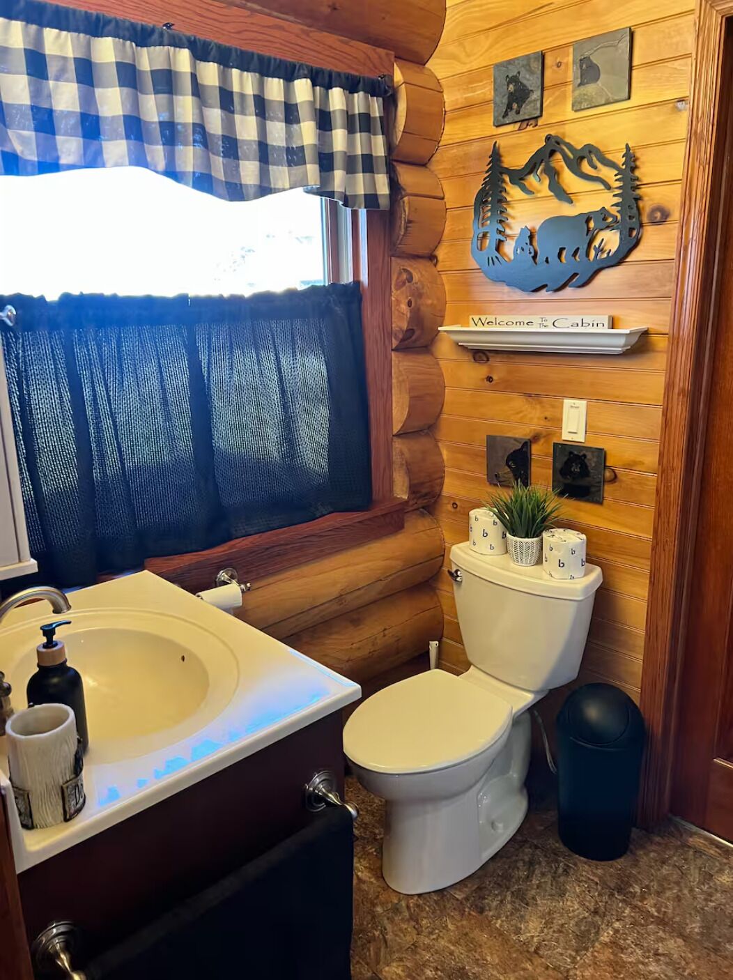 Une salle de bain de style rustique, avec un mur en bois, un lambrequin à carreaux à la fenêtre, un lavabo avec un miroir, des toilettes et une étagère décorée sur le thème des ours.