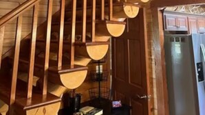 Eine Holztreppe mit Geländer, einer Deckenleuchte und Blick in einen Küchenbereich mit Schränken und einem Kühlschrank.