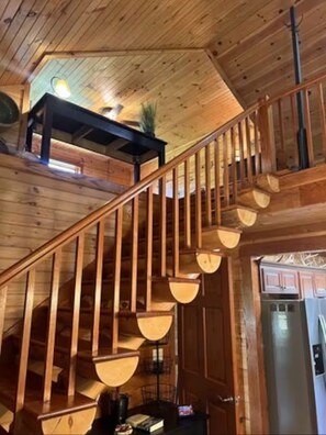 Eine Holztreppe mit Geländer, einer Deckenleuchte und Blick in einen Küchenbereich mit Schränken und einem Kühlschrank.