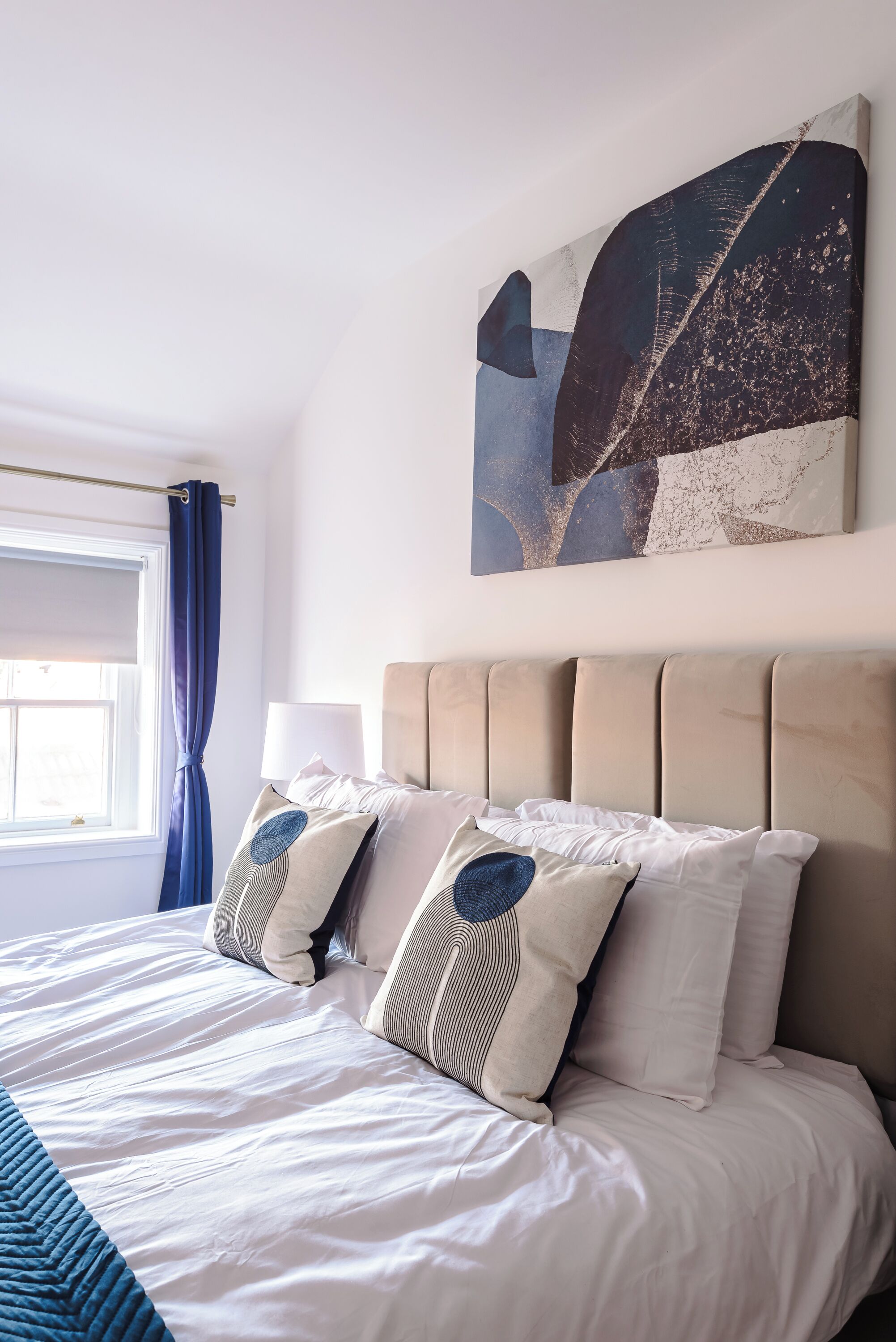 A neatly made bed with a headboard, a window with a curtain, and a wall-mounted abstract painting.