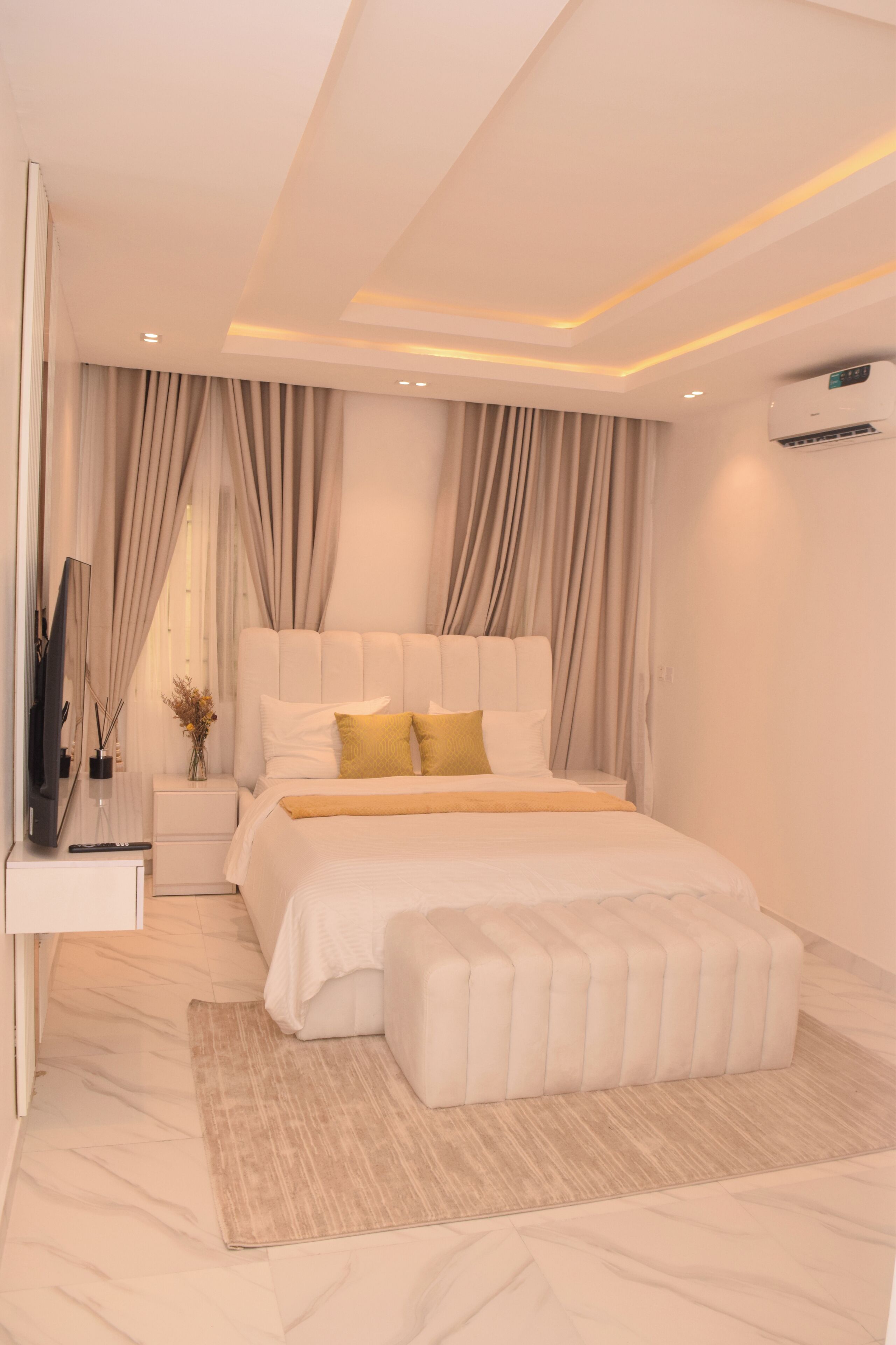 A modern bedroom with a large bed, a bedside table, a TV, and an air conditioning unit.