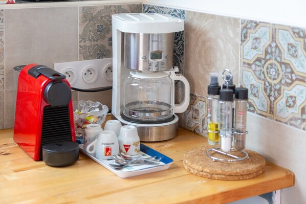 A coffee maker, a red electric kettle, a tray with cups and condiments, and a cork coaster on a wooden countertop.