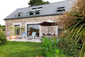 A stone house with a sloped roof, a patio with lounge chairs, and a large umbrella.