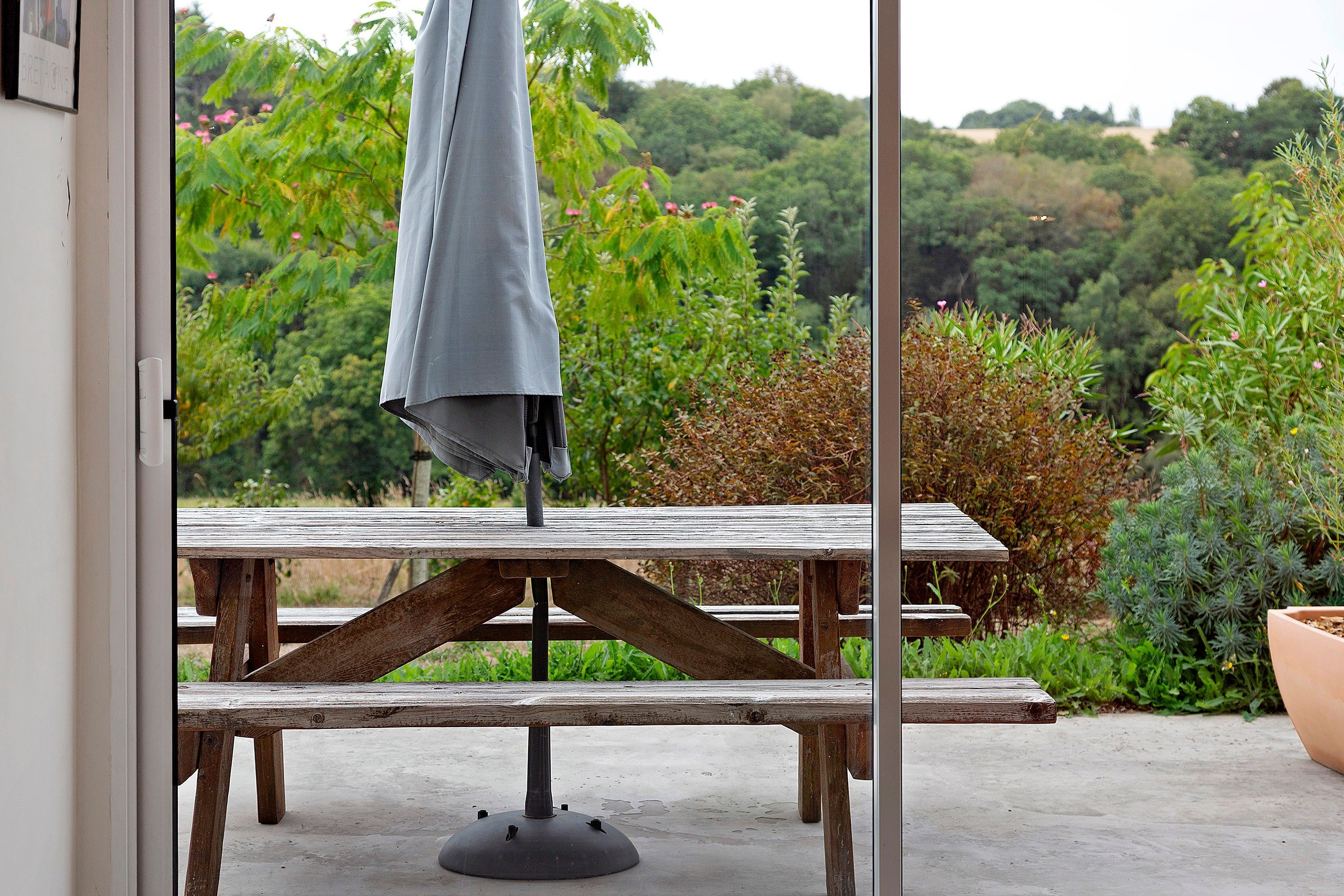 Una mesa de picnic de madera con base metálica, un paraguas y vistas a un jardín con diversas plantas y árboles.