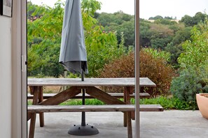 A wooden picnic table with a metal base, an umbrella, and a view of a garden with various plants and trees.
